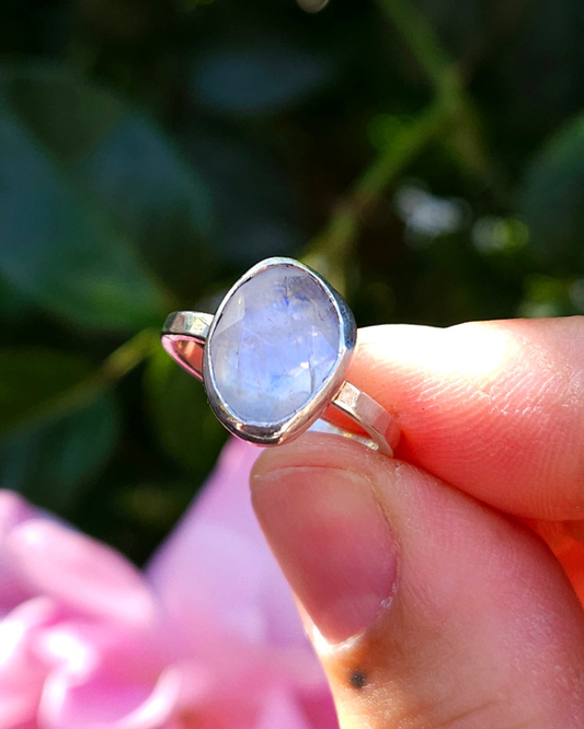 A rose-cut rainbow moonstone and .925 silver ring are held between finger and thumb with green foliage and pink flowers in the background. The responsibly sourced stone is freeform and displays beautiful, otherwordly, luminescence throughout. 