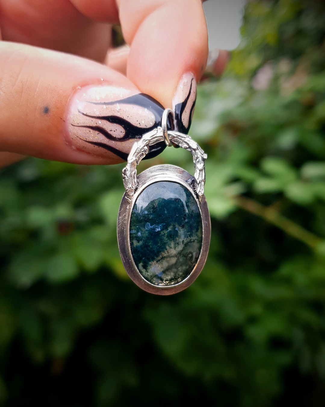 A rich, green, translucent moss agate sits in an oxidised silver bezel. The bail loop of the pendant is a highly textured casting of a twig in solid silver. The pendant is held up by two fingers with long painted nails with black fire details. The background is a blurred, deep emerald green bush