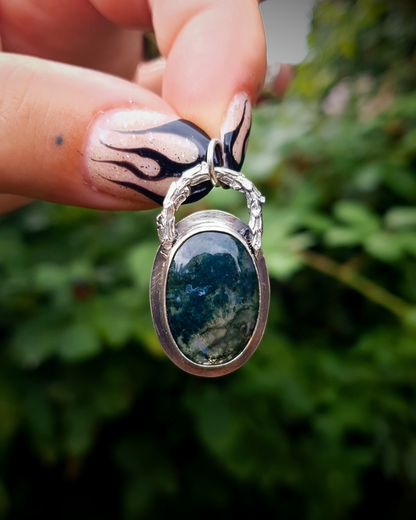 A rich, green, translucent moss agate sits in an oxidised silver bezel. The bail loop of the pendant is a highly textured casting of a twig in solid silver. The pendant is held up by two fingers with long painted nails with black fire details. The background is a blurred, deep emerald green bush