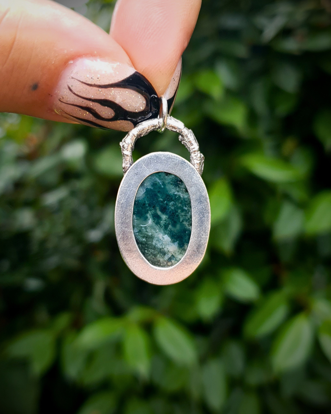 The back view of a green and silver pendant is showing. The silver backing has a window-like cut-out to let the light pass through a rich, green, translucent moss agate which sits in an oxidised silver bezel. The bail loop of the pendant is a highly textured casting of a twig in solid silver. The pendant is held up by two fingers with long painted nails with black fire details. The background is a blurred, deep emerald green bush
