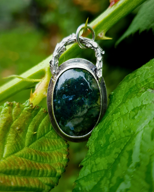 A rich, green, translucent moss agate sits in an oxidised silver bezel. The bail loop of the pendant is a highly textured casting of a twig in solid silver. The pendant is lightly speckled in rain drops and photographed amongst the leaves and twigs of a bramble bush. The whole image is luscious green and bathed in natural light.