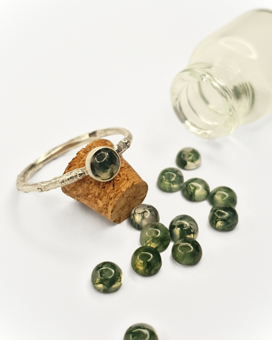 A little, glass bottle lays on its side with its content spilled out over a white background. The contents are 12 little moss agate gemstones. A beautiful, twig-textured, sterling silver ring with a moss agate setting rests against the cork stopper of the class bottle.