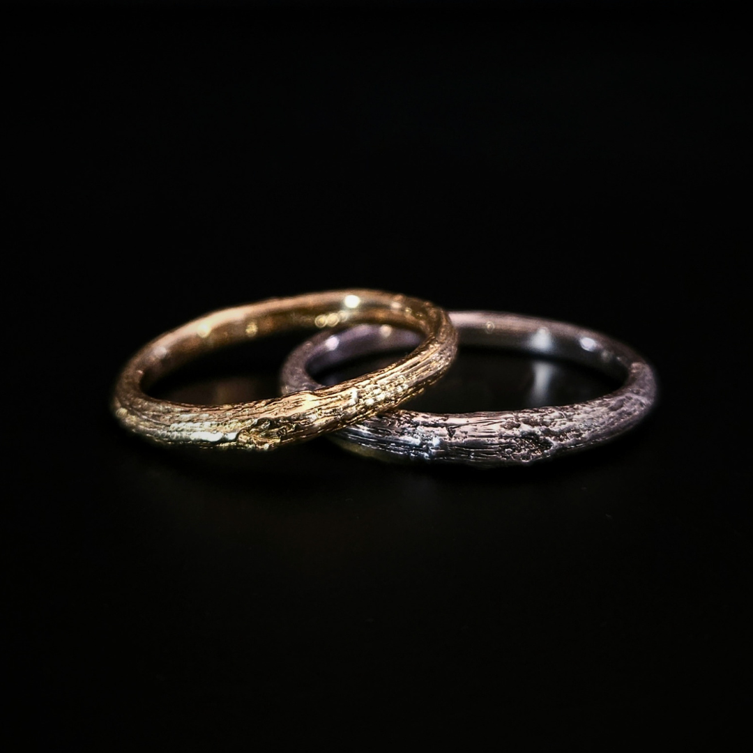 Two organic, oak-bark textured rings, one in recycled 9 carat gold and one in recycled sterling silver, overlapping on a solid black background.  The light plays in the natural ridges and texture of the wood-like surface.