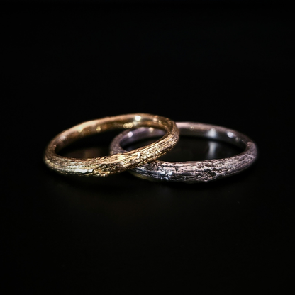 Two organic, oak-bark textured rings, one in recycled 9 carat gold and one in recycled sterling silver, overlapping on a solid black background.  The light plays in the natural ridges and texture of the wood-like surface.