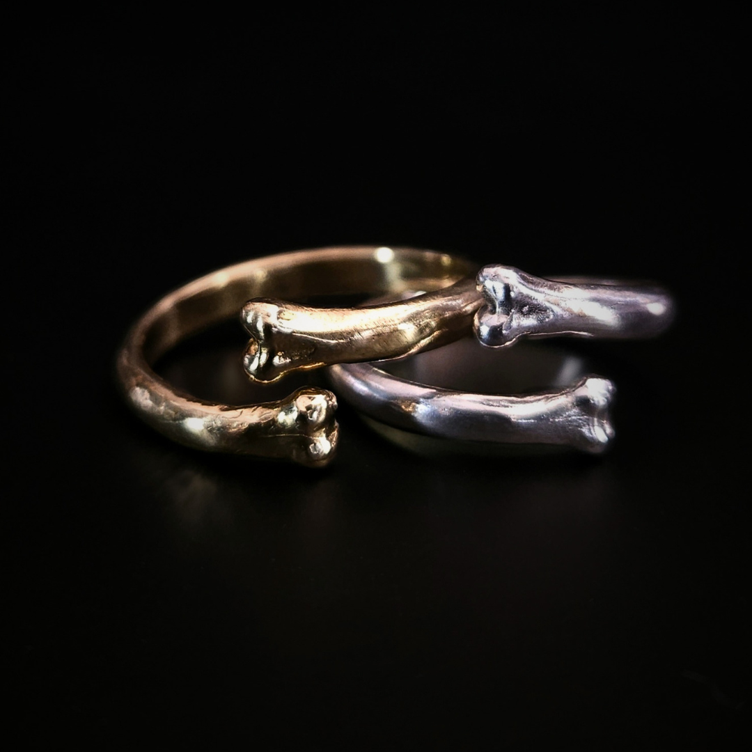 Two open style rings with bone ends sit on a black background. They slightly overlap. The one on the left is made of yellow gold and the one on the right is made of sterling silver. Both reflect the light in a soft glow.
