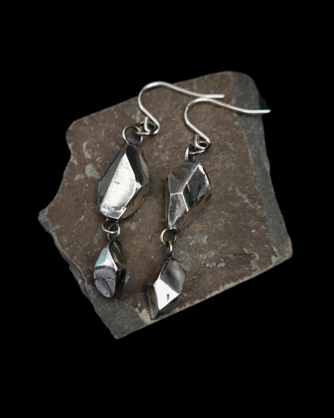 A pair of silver drop hook earrings with faceted, geometric sterling silver charms, displayed on an angular piece of slate against a solid black background.