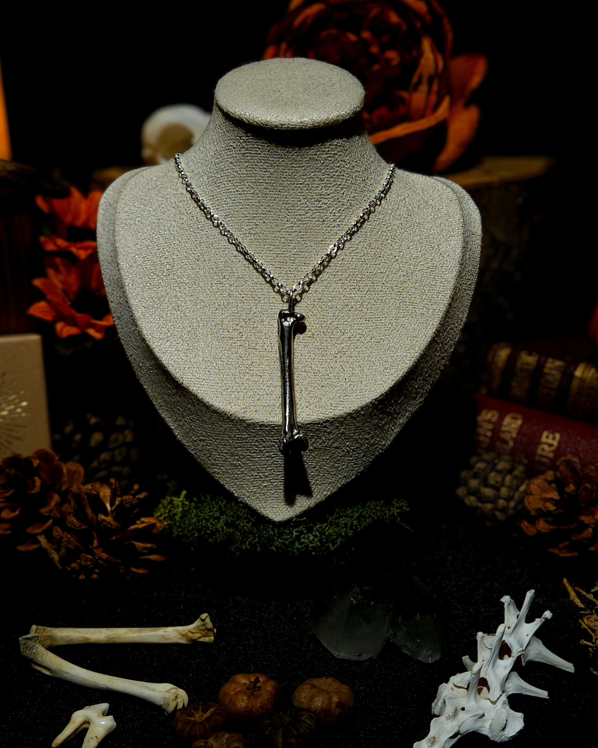 A large solid silver foraged animal bone pendant on a silver chain displayed on a beige vintage style jewellery bust. The photograph is styled in a witchy, moody theme with vintage red books, candles, crystals, teeth , bones and autumnal botanicals.