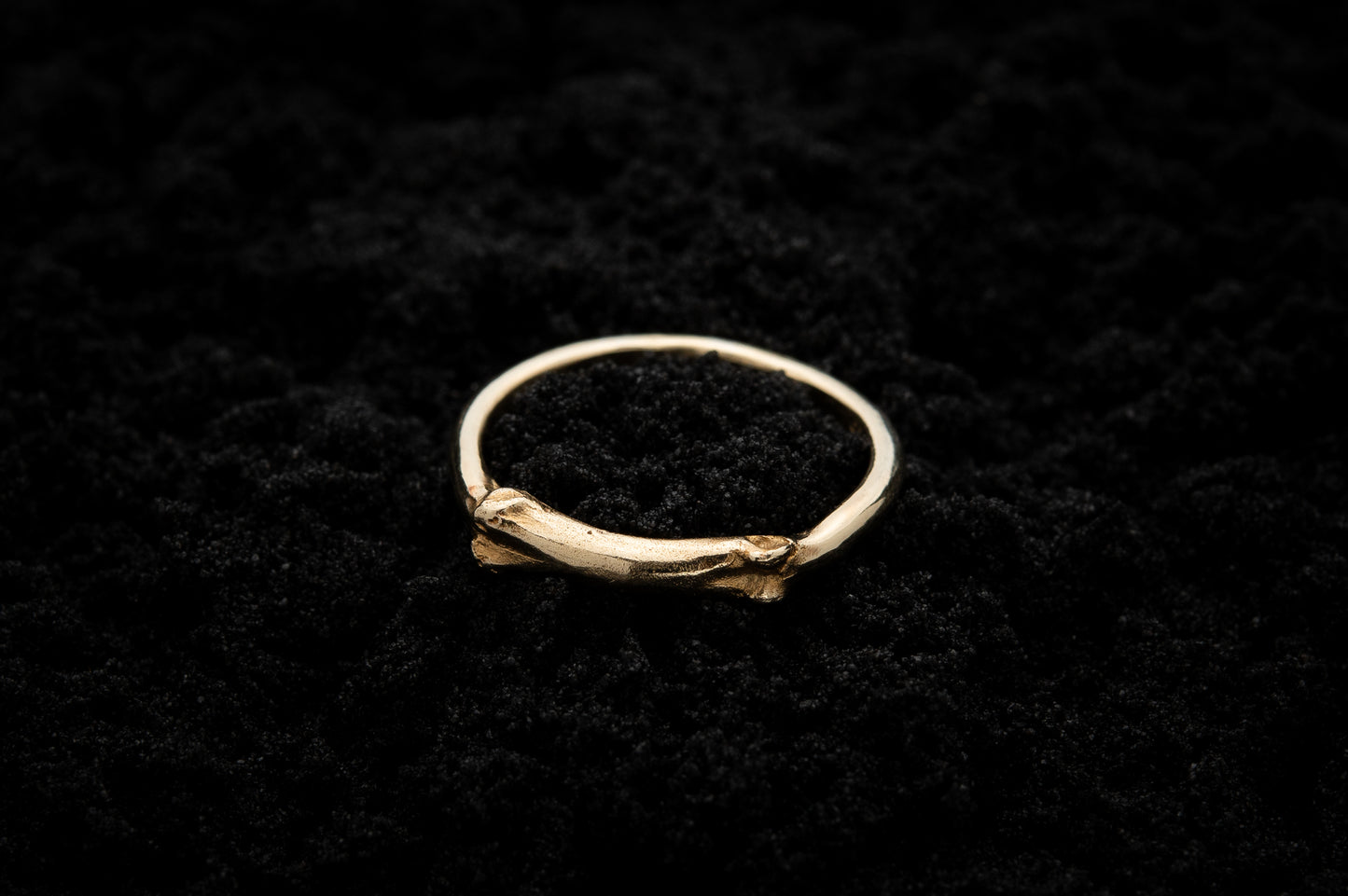 9ct gold ring ring lays on black sand. Part of the band of the ring is the solid gold casting of a little bone