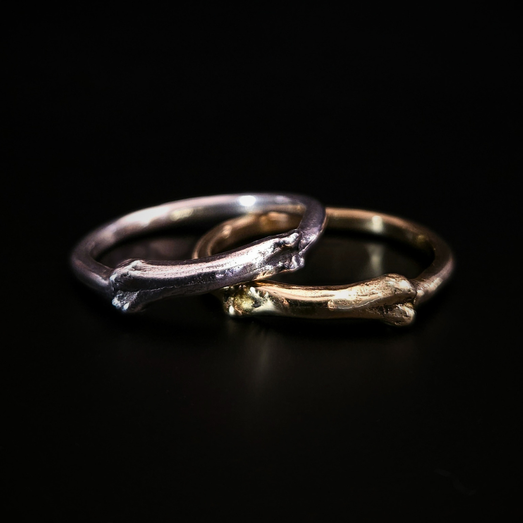 Two gothic rings with a small bone at the front sit on a black background, overlapping. The one on the left is solid silver and sits over the top of the 9 carat yellow gold one on the left.