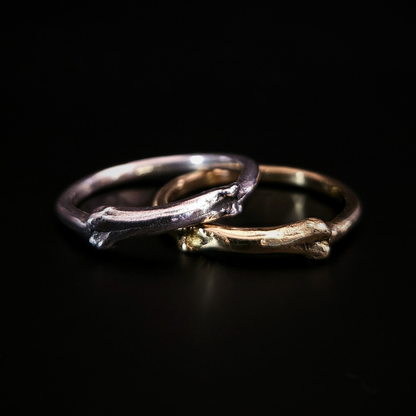 Two gothic rings with a small bone at the front sit on a black background, overlapping. The one on the left is solid silver and sits over the top of the 9 carat yellow gold one on the left.