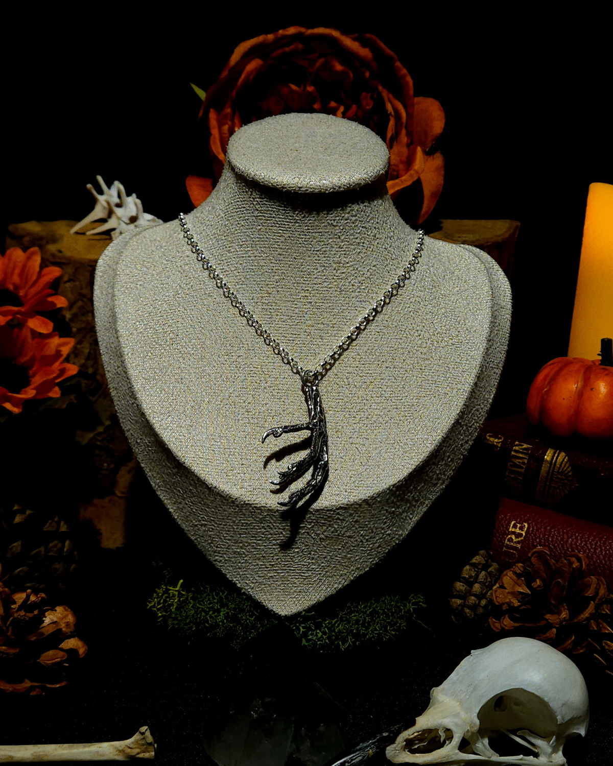 A solid .925 silver  pigeon claw pendant displayed on a vintage style jewellery bust. The image is styled in an autumnal, folklore inspired, moody setting with candles, pumpkins, bones, and warm toned botanicals around it the bust in the centre.