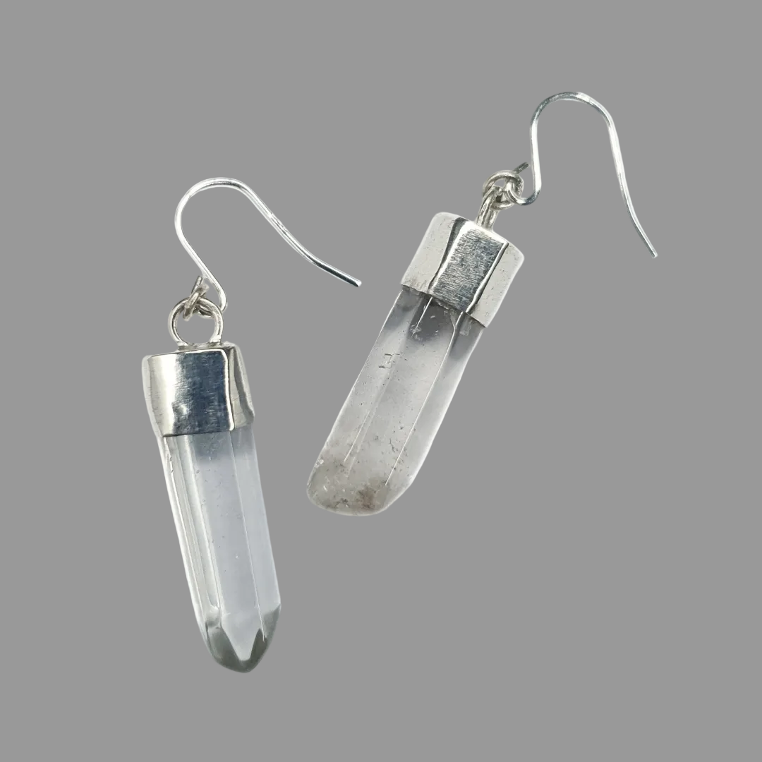 A pair of quartz and silver earrings are displayed on a grey background.
They are comprised of a clear quartz point coming from a handmade silver setting with a silver hook. 