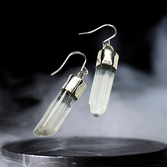 A pair of quartz and silver earrings are displayed on a black smoky background.
They are comprised of a clear quartz point coming from a handmade silver setting with a silver hook. 