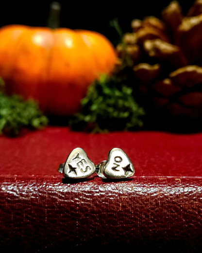 A pair of small, .925 silver ouija planchette stud earrings sit on a vintage red book with autumnal decor, like a halloween pumpkin, moss & pinecone, out of focus in the background. The earring on the left says "YES" and the one on the right says "NO" and they both have a 4-pointed star at the bottom of each pointer.