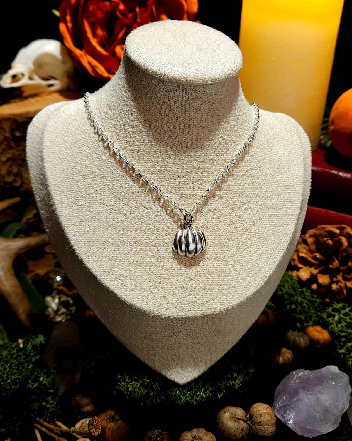 A silver chain with a solid silver halloween pumpkin pendant displayed on a vintage beige jewellery bust. The picture is themed in a warm, dark, moody setting with candles, pumpkins, bones, and witchy, autumnal botanicals around it.