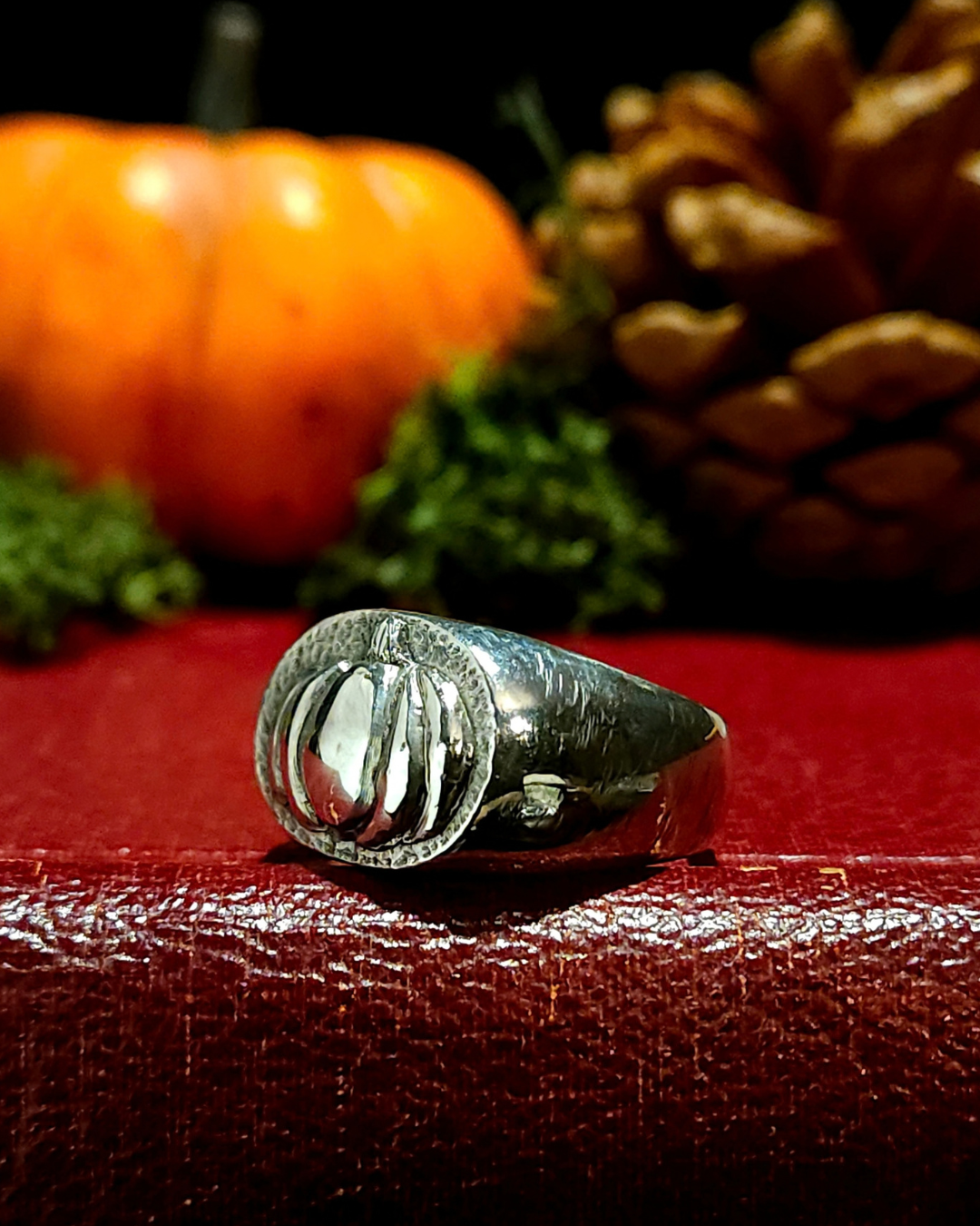 A sterling silver signet ring with 3D pumpkin detailing, displayed on a textured vintage red book with a moss, an autumnal pumpkin and pine cone in the background out of focus.