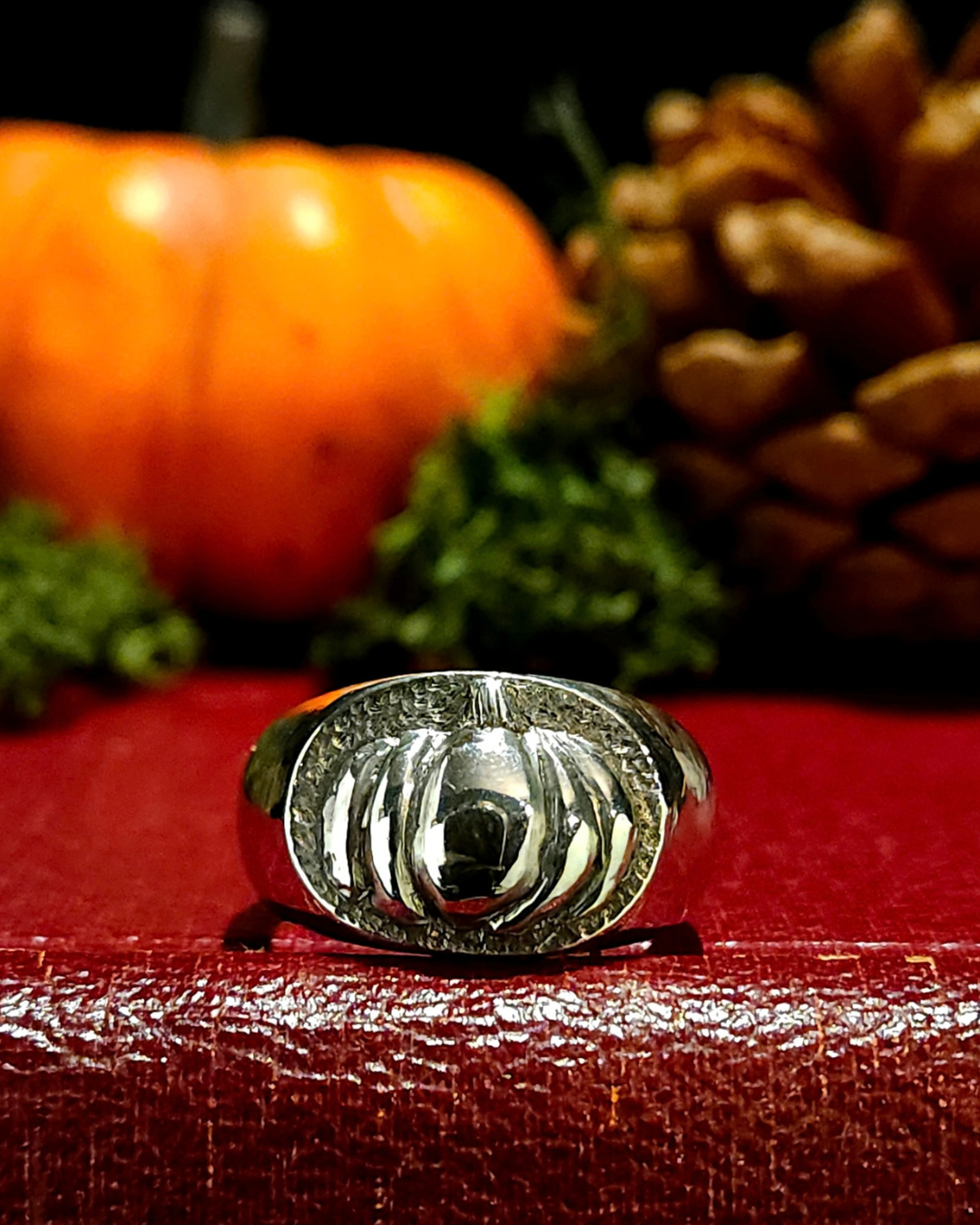A chunky .925 silver signet ring with 3D pumpkin detailing, displayed on a textured vintage red book. It is oxidised to reveal detailing and polished back to a nice shine. The background is themed for autumn and halloween with a moss, an orange pumpkin and pine cone - all out of focus.
