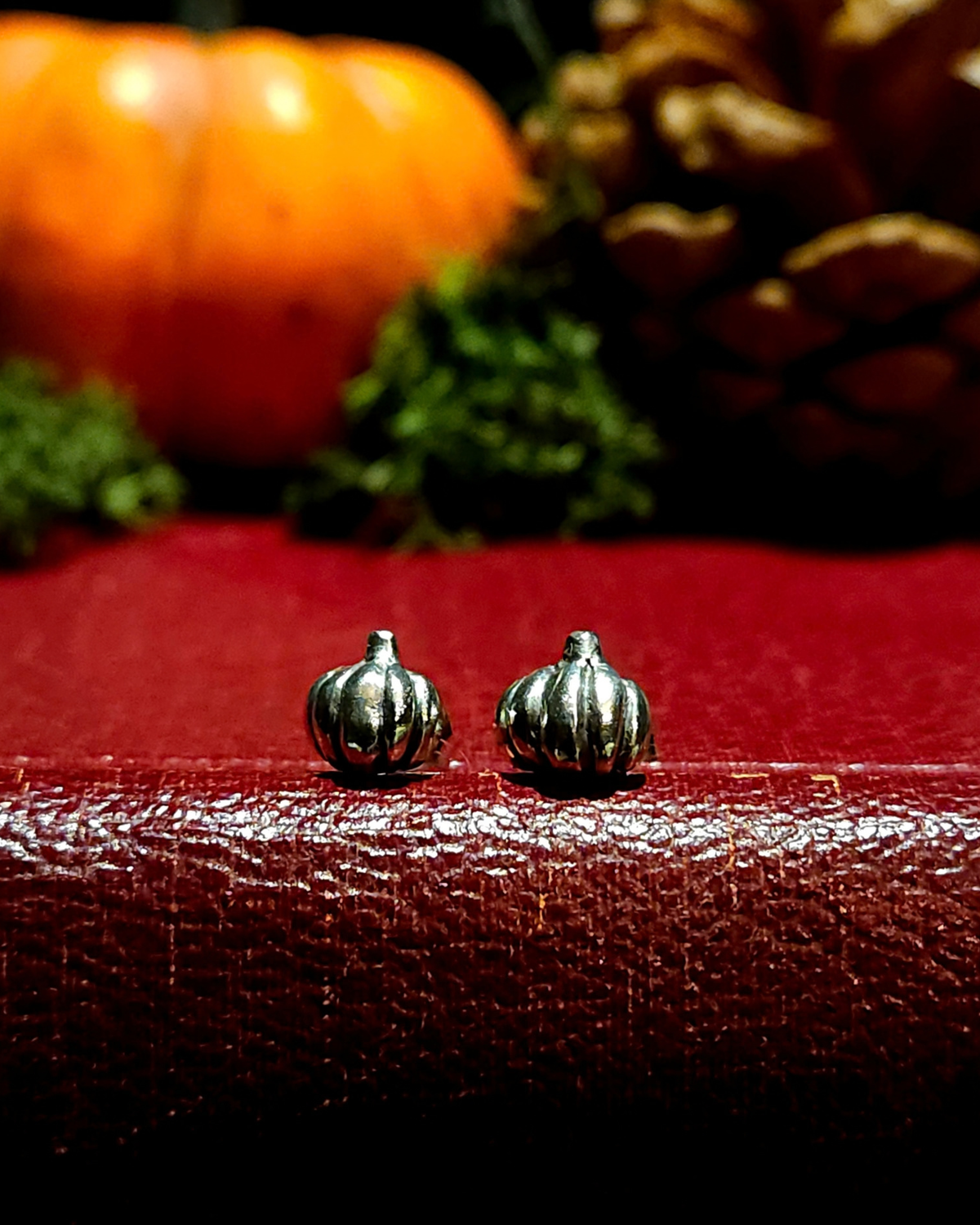 A pair of small, .925 silver pumpkin stud earrings sit on a vintage red book with autumnal decor, like a halloween pumpkin, moss & pinecone, out of focus in the background