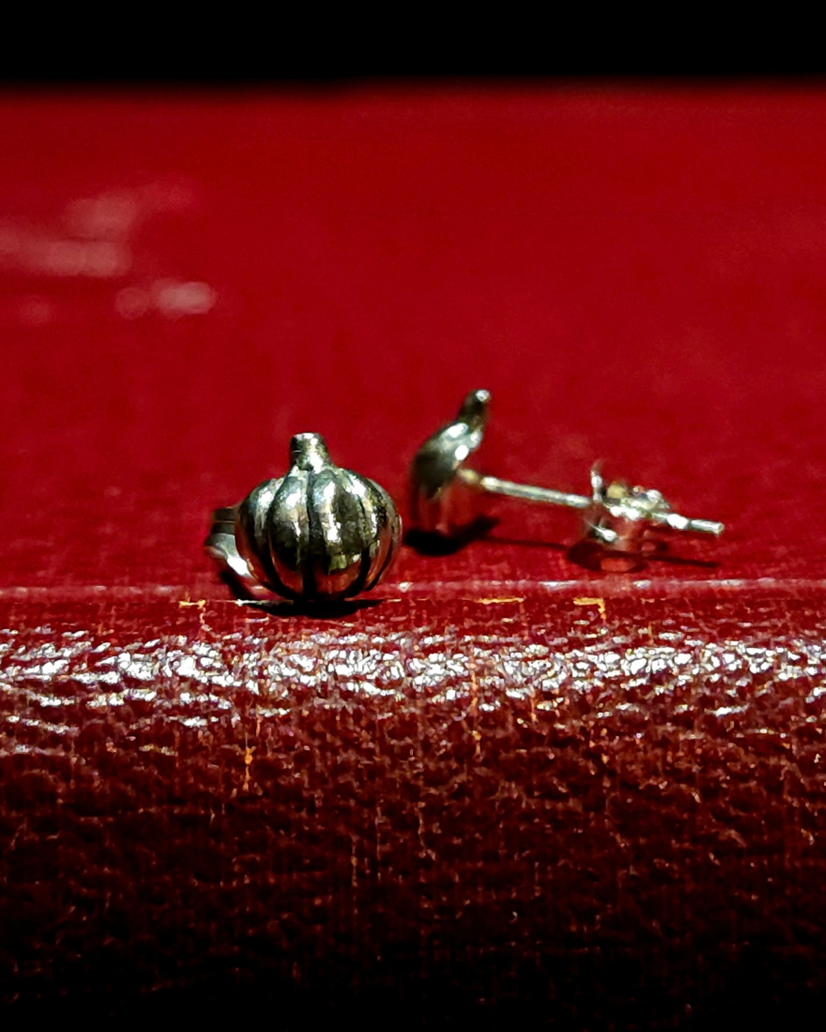 A pair of sterling  silver pumpkin stud earrings sit on a vintage red book. The one on the right is facing forward and in focus, the one on the right is out of focus, facing sideways to show its flat back and butterfly fastener.