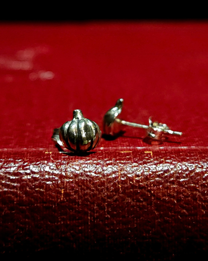 A pair of sterling  silver pumpkin stud earrings sit on a vintage red book. The one on the right is facing forward and in focus, the one on the right is out of focus, facing sideways to show its flat back and butterfly fastener.