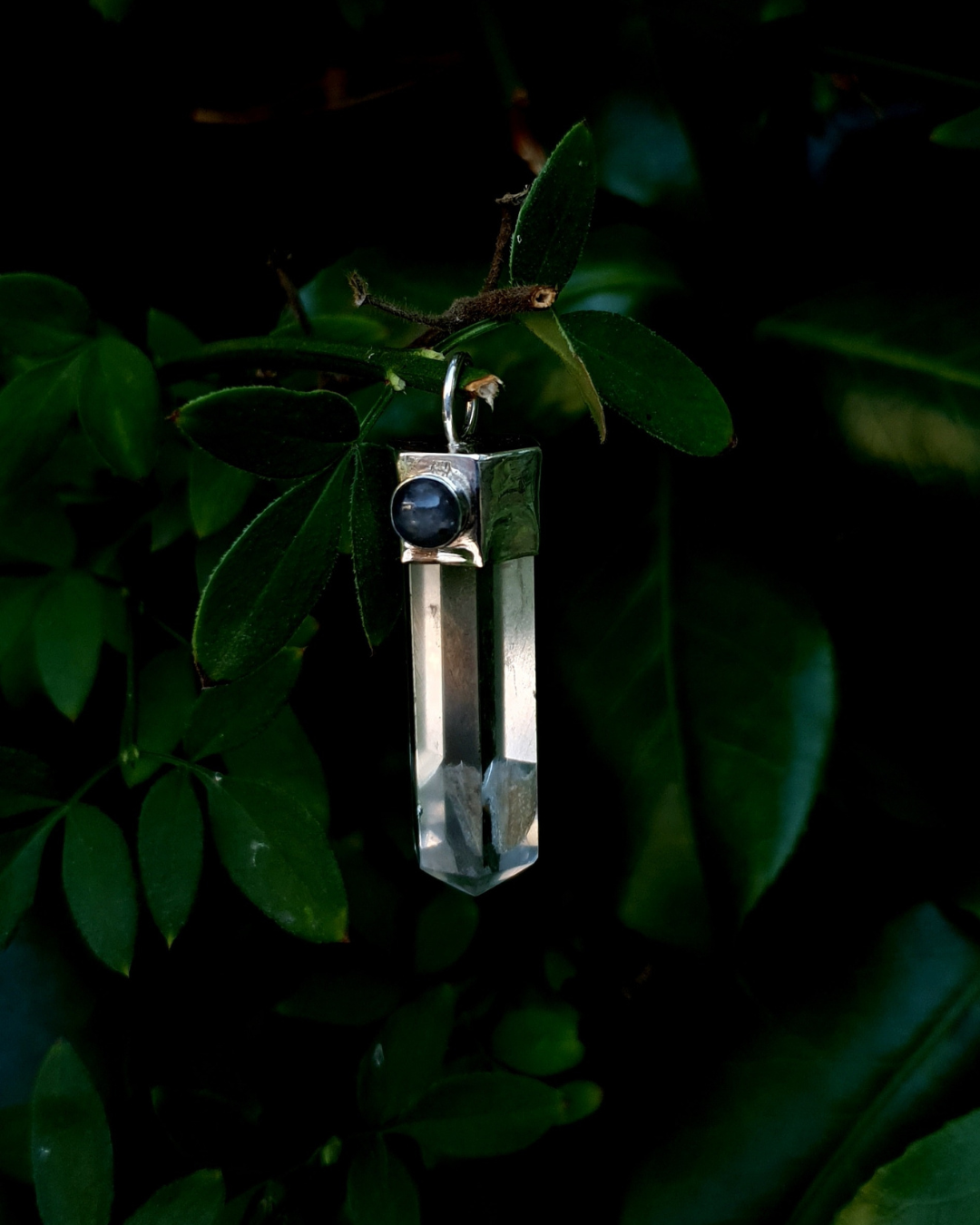A pendant hangs from a branch surrounding by gorgeous green leaves. The pendant comprises of a clear quartz point in a sterling silver setting. A 5mm round labradorite adorns the silver setting and is illuminated by the daylight. There is a large, beautiful inclusion at the bottom of the quartz point.