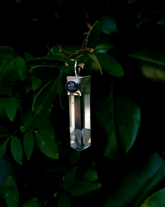 A pendant hangs from a branch surrounding by gorgeous green leaves. The pendant comprises of a clear quartz point in a sterling silver setting. A 5mm round labradorite adorns the silver setting and is illuminated by the daylight. There is a large, beautiful inclusion at the bottom of the quartz point.