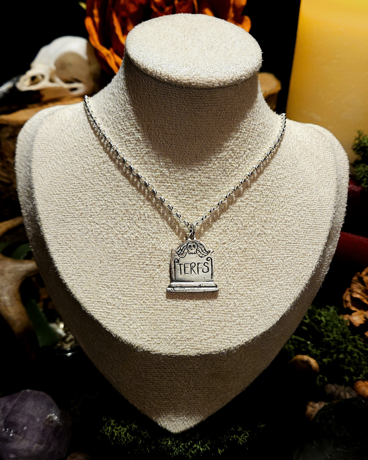 A sterling silver tombstone pendant featuring a winged skull, engraved with 'TERFS', is displayed on a beige vintage style bust. The bust is set against a dark, mystical background with candles, a skull, crystals and other macabre and dark academia themed items