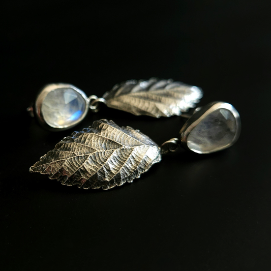 A pair of botanical styled, real silver & rainbow moonstone earrings lay on a black background.
The focus of the image is on the highly textured leaves, which hang from the gemstones. They are .999 silver bramble leaves containing a high level of natural detail including a natural curl and depth along with intricate veining.
