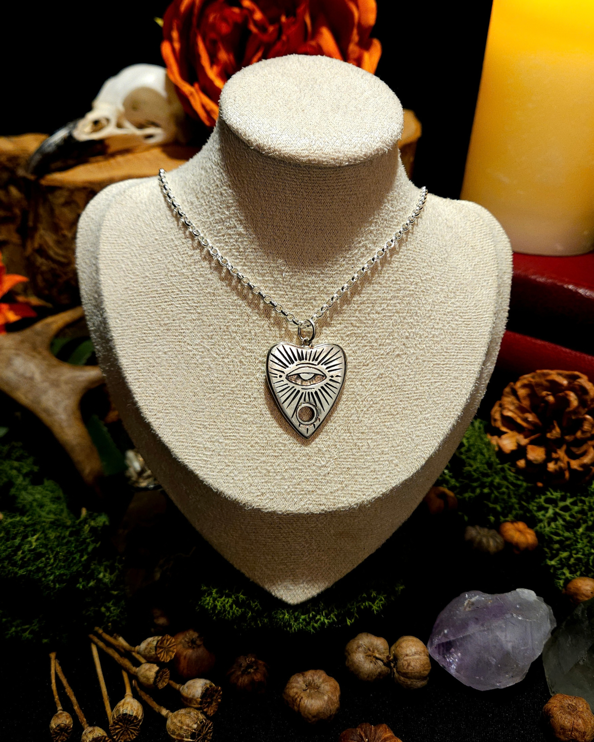 A silver ouija planchette pendant necklace with an eye of providence design, displayed on a textured vintage style velvet jewelry bust. The picture is styled with mystical props including a crow skull, amber flowers, a crystal, deer antlers, pinecones, and a candle in the background.