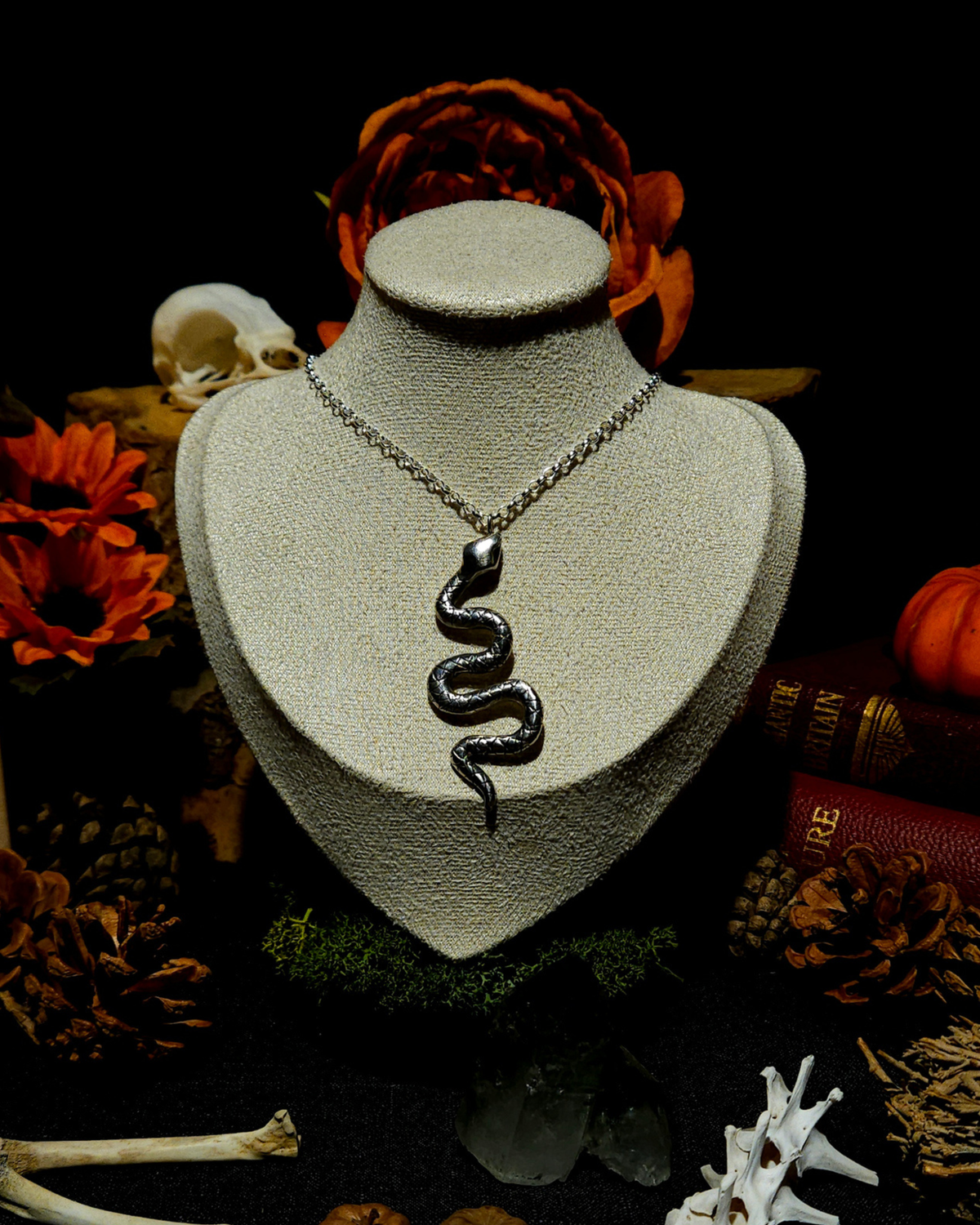 An extra large solid .925 silver snake pendant displayed on a vintage style jewellery bust. The image is styled in an autumnal, witchcraft inspired, moody setting with candles, pumpkins, bones, and warm toned botanicals around it the bust in the centre.