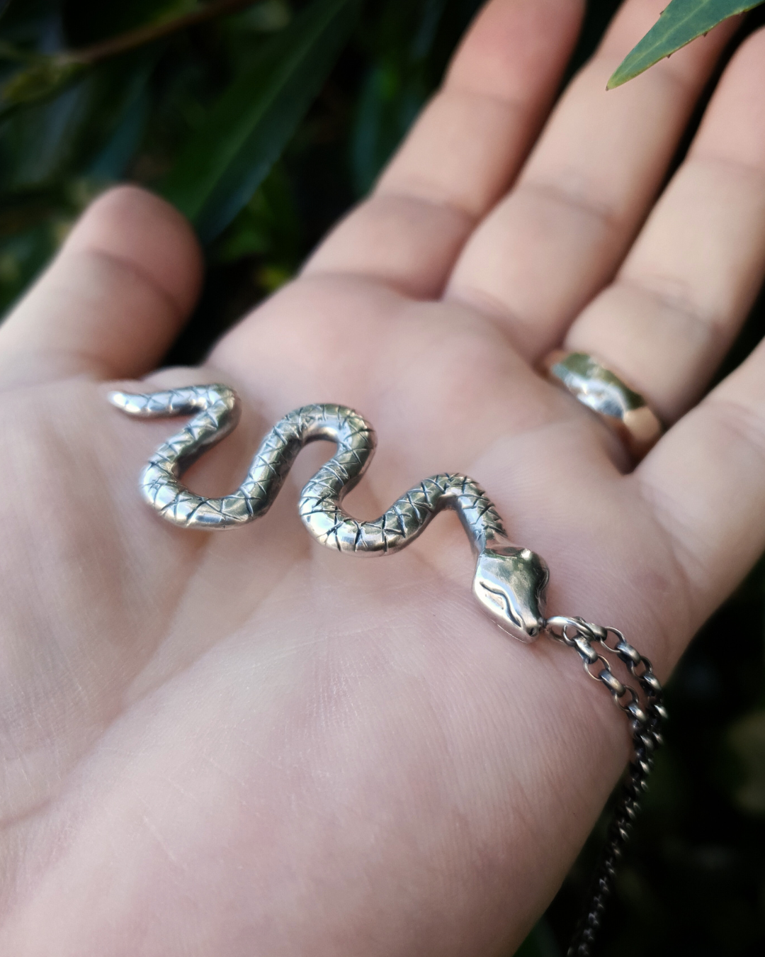 A large, solid sterling silver snake pendant sits on an open palm with a natural backdrop of dark green leaves.
The snake is in a wavy position with its head facing towards the camera and the chain is attached to a jump ring on its head.