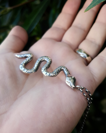 A large, solid sterling silver snake pendant sits on an open palm with a natural backdrop of dark green leaves.
The snake is in a wavy position with its head facing towards the camera and the chain is attached to a jump ring on its head.