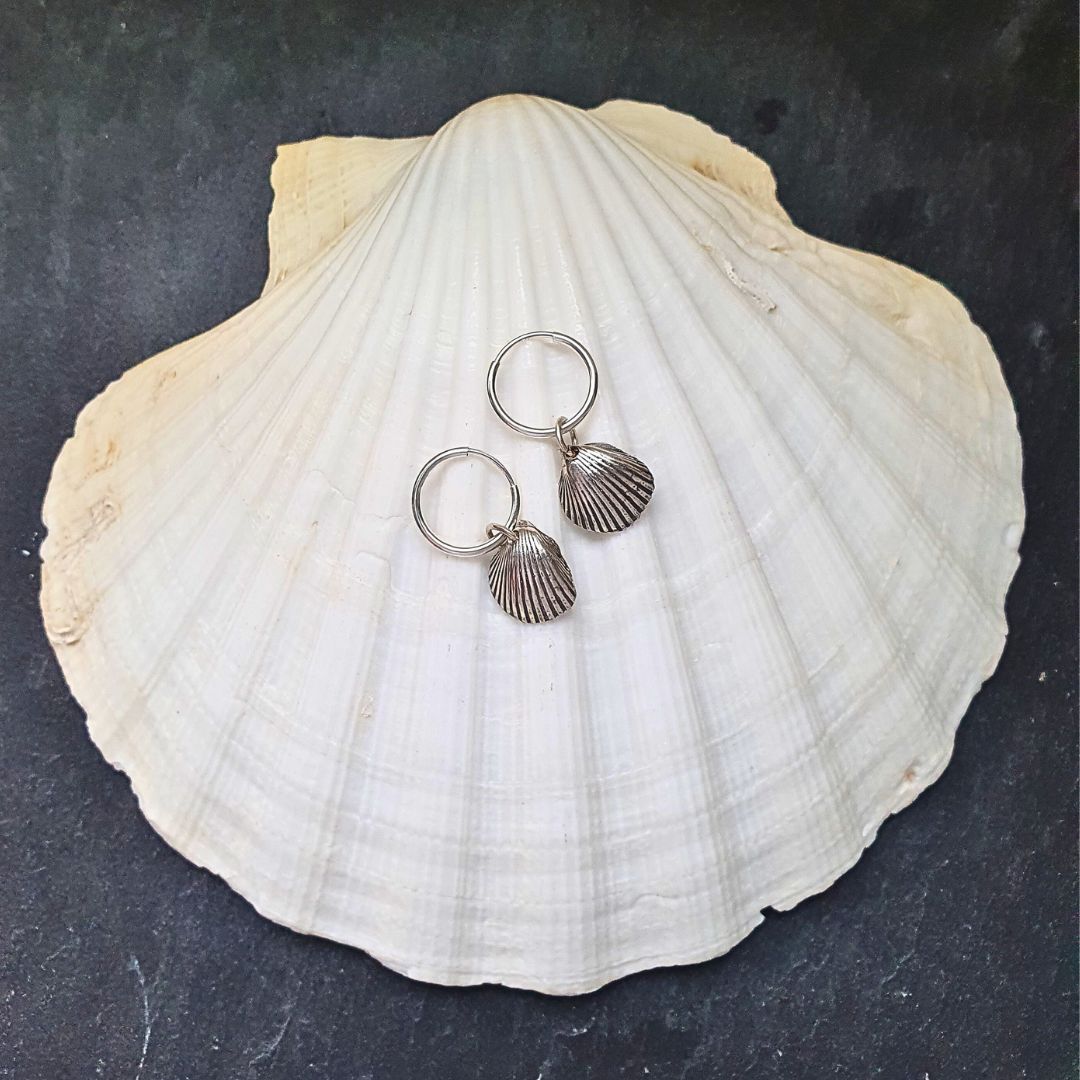 A pair of dainty, .999 fine silver shell hoop-earrings sit on a large white clam shell