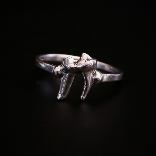 A small, alternative style, solid silver ring made using the casting of a real baby fox tooth, sits against the black background, photographed from the front. The pale silver ring has been oxidised to show the natural wear and texture of the foxes tooth from its life in the wild. It perfect for fans of a witchy and gothic aesthetic and fans of taxidermy and oddities