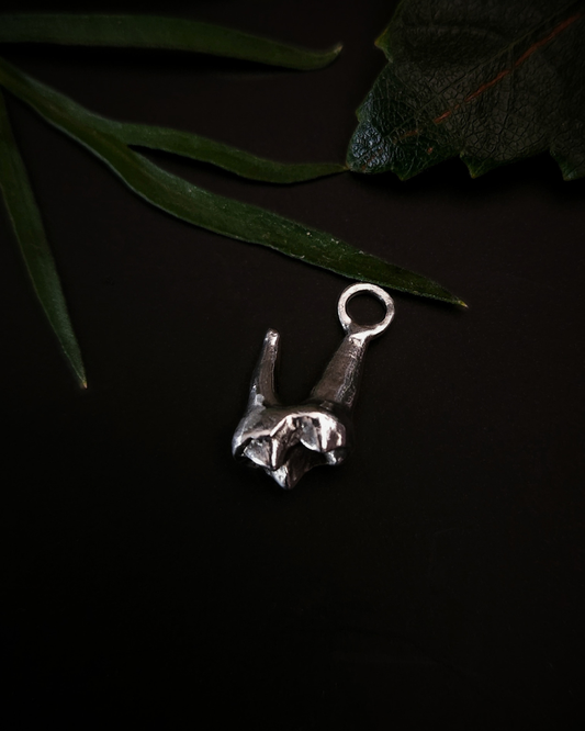 a small, silver pendant made from the casting of a baby fox tooth sits on a black background with a few pieces of green foliage around it. The photograph is taken from the top down. This is a dainty taxidermy style pendant for fans of gothic jewellery, the macabre, unusual and taxidermy.