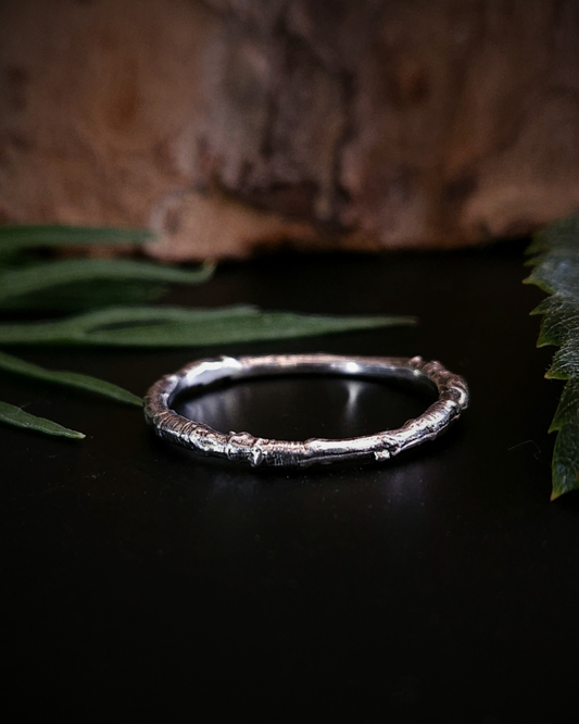 A sterling silver ring made from the casting of a thin twig sits on a black surface with some brown bark and green foliage out-of-focus in the background. The inside of the ring is nice and smooth for comfort and the light reflects from it in a soft pale glow.