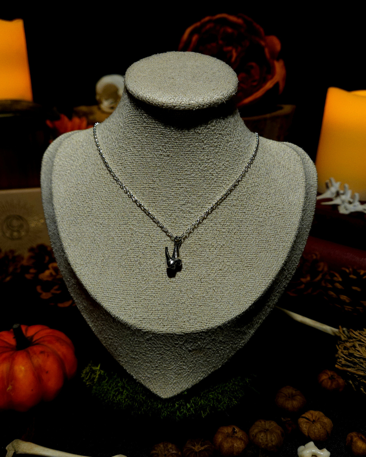 A silver chain with a solid silver fox tooth pendant displayed on a vintage beige jewellery bust. The picture is themed in an atmospheric, moody setting with candles, pumpkins, bones, and witchy, autumnal botanicals around it.