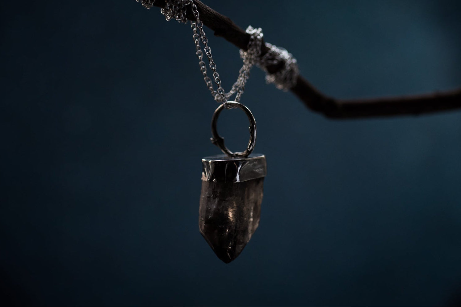 a smoky quartz crystal point with twig detailing made from solid silver hangs on a silver chain. the chain is wrapped around a twig hanging against a night sky.