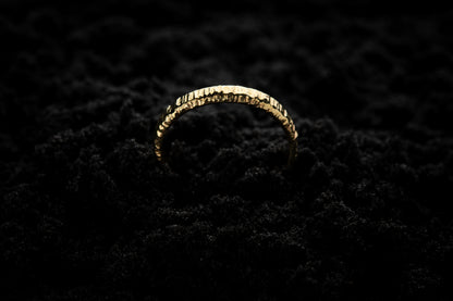 A slim, yellow gold wedding ring sits in black sand to contrast its warm, luscious glow. There are hammered striations around the band like the suns rays. It is half submerged in the sand and breaks through the surface like a golden rainbow.