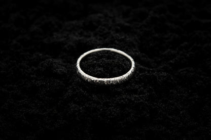 A slim, silver wedding ring sits atop black sand to contrast its bright, shiny glow. There are hammered striations around the band like the suns rays.