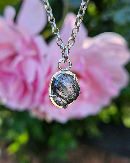 A sterling silver and gemstone pendant hangs in front of an out-of-focus, pink rose bush. The gemstone is a freeform rose-cut tourmalinated quartz. The sun shines thorough the back, illuminating the quartz and showing the black included striations of tourmaline. The tourmaline is denser on the outer edges, seeming to reach for eachother across the centre of the stone.