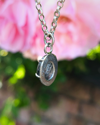 .925 silver pendant in front of pink flowers. The pendant is hanging so you can see the smooth back with a cut-out to allow the light through  illuminate the stone from within.