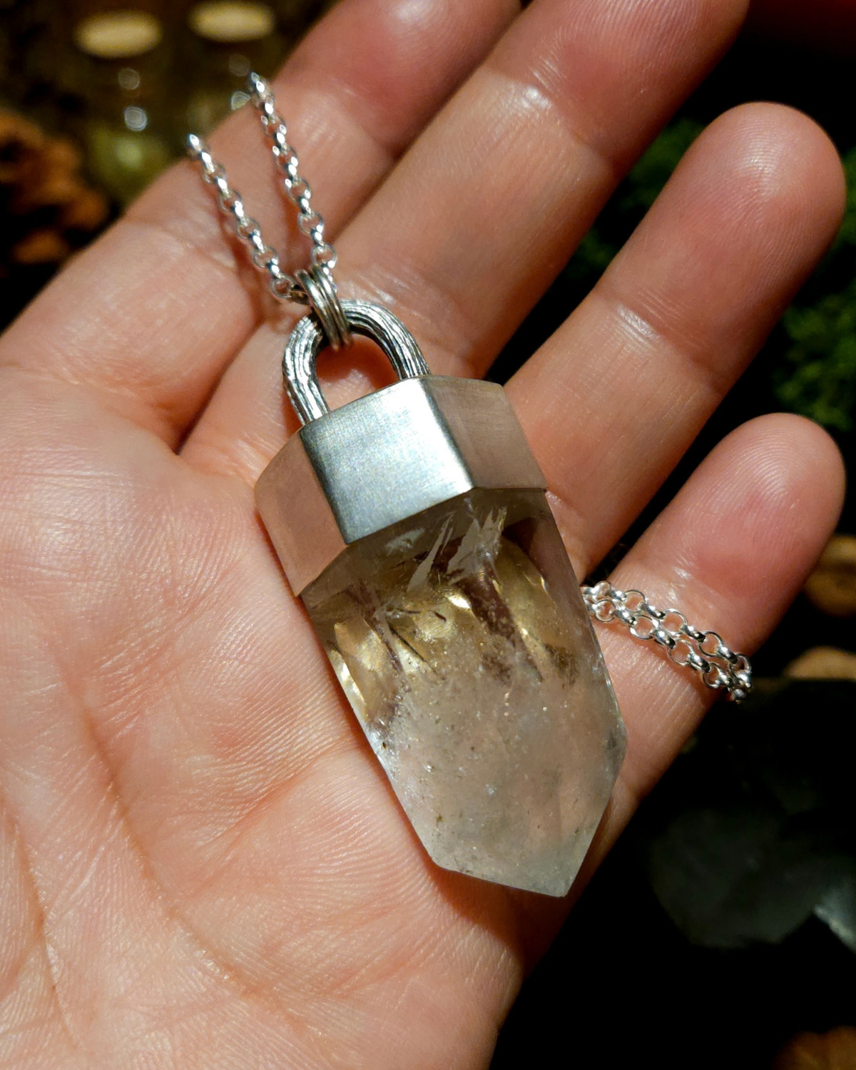 A large quartz point pendant made from solis silver, featuring an oak twig bail, is held in an open palm. The photograph is taken from above and there is a witchy, dark academia themed background out of focus beneath the hand.
