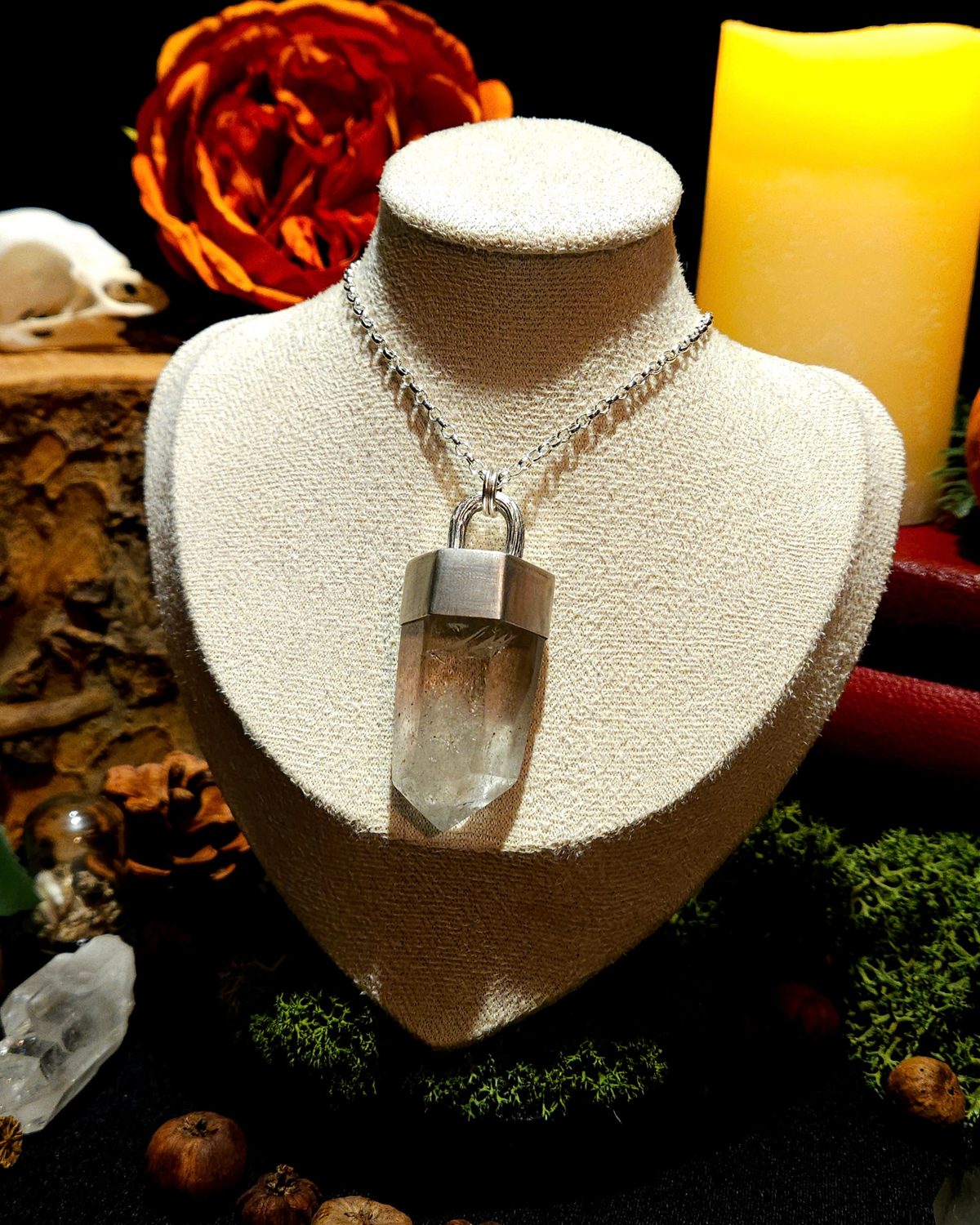 A large quartz point pendant on a thick silver chain is displayed on a vintage beige jewellery bust. The picture is themed in an atmospheric, moody setting with candles, bones, and witchy, autumnal botanicals surrounding the bust.