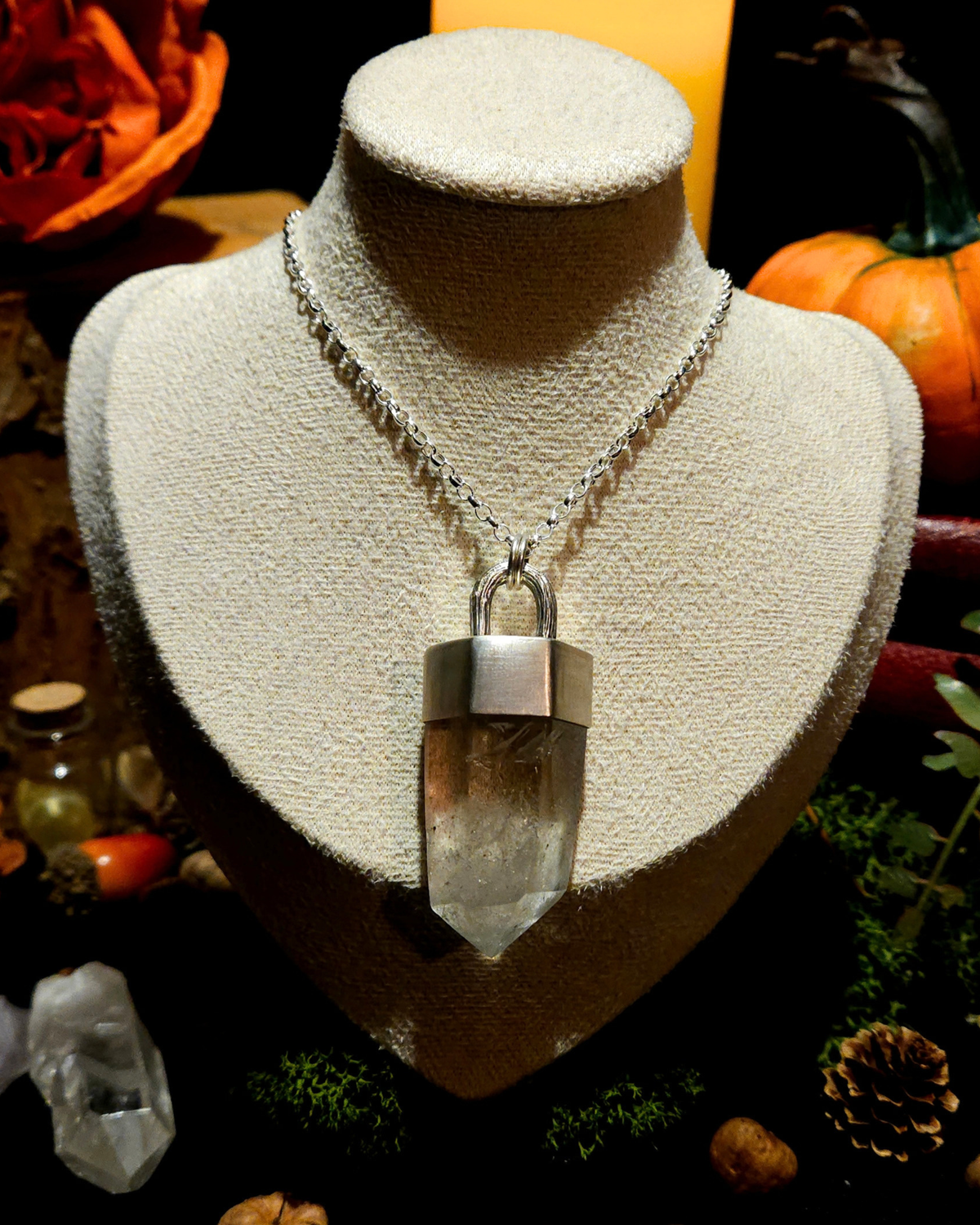 A large sterling silver & quartz point pendant on a thick silver chain is displayed on a vintage beige jewellery bust. The picture is themed in an autumnal, moody setting with candles, a pumpkin, bones, and witchy, autumnal botanicals surrounding the bust.