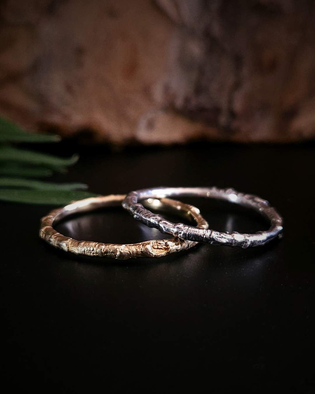 2 rings made from the castings of a thin twig sit on a black surface with an out-of-focus natural background. The one on the left is made from yellow gold and the one on the right is made from sterling silver. inside the rings are nice and smooth for comfort and the light reflects from it in a soft glow.
