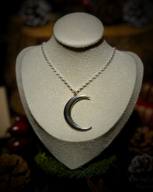 A silver chain necklace with a large sterling silver crescent moon pendant displayed on a cream jewellery bust. The photograph is styled in a autumnal nature theme with acorns, pine cones, moss & tree bark out of focus in the background