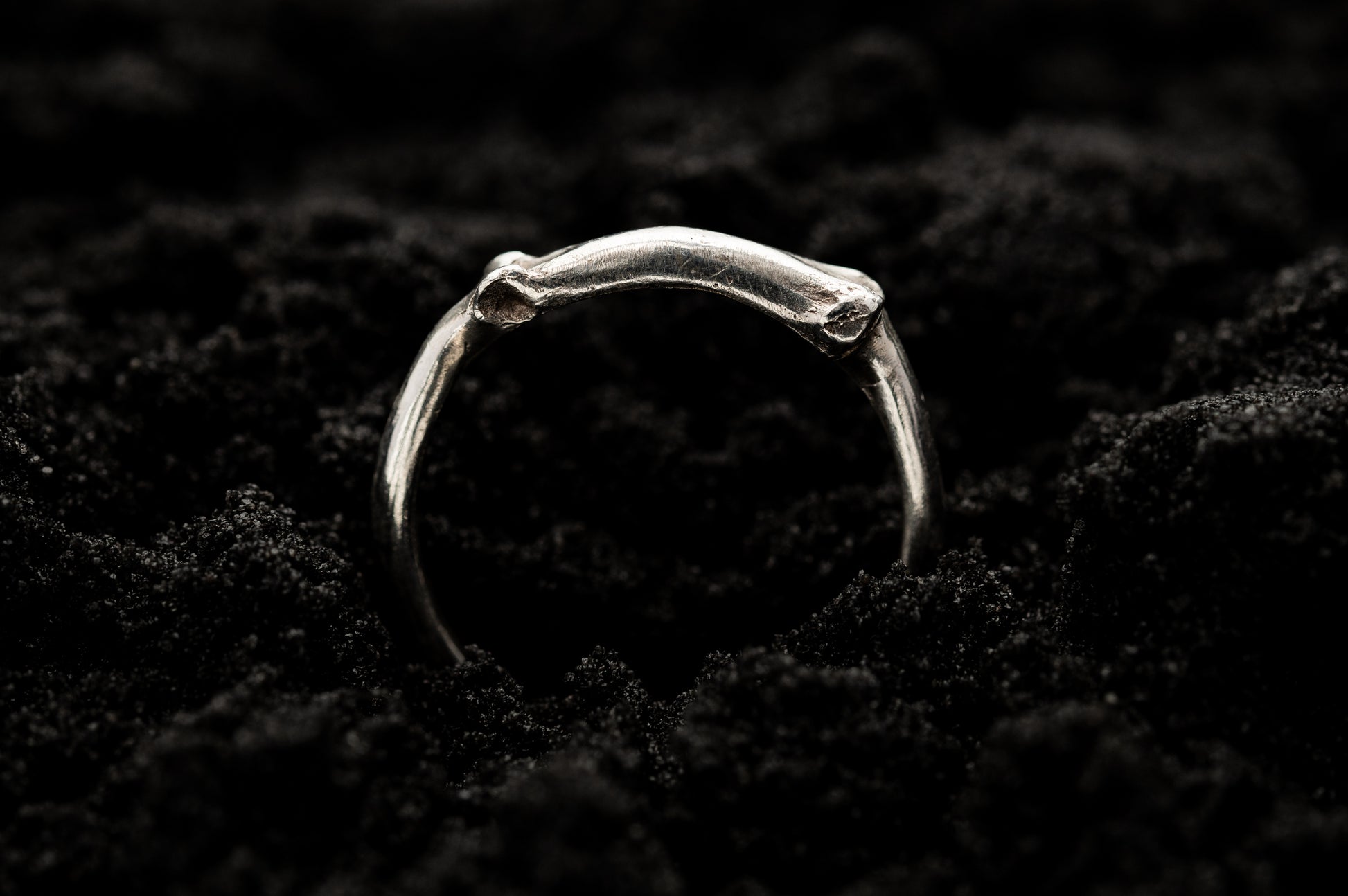 A sterling silver ring sticks out of black sand in bright contrast. Part of the band of the ring is the solid silver casting of a little bone