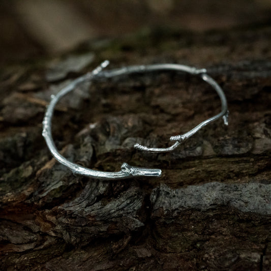 A sterling silver twig bracelet sits on the dark bark of a fallen tree.