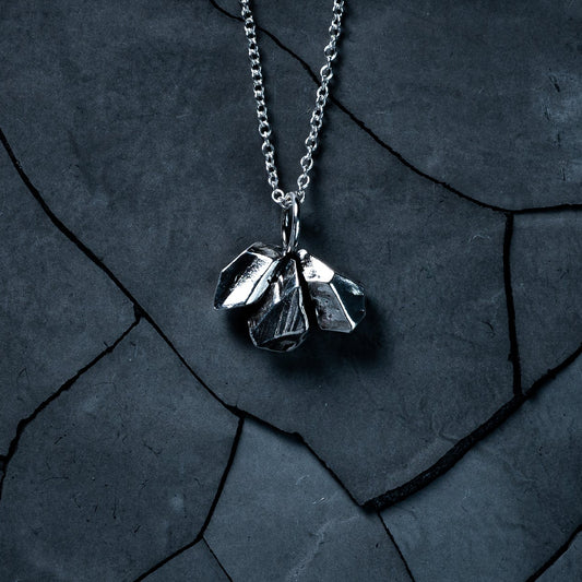 A sterling silver necklace resting on a dark, cracked stone surface. The pendant is made of a cluster of 3 rough-cut nuggets of solid silver. Geometric, unisex, minimalist.